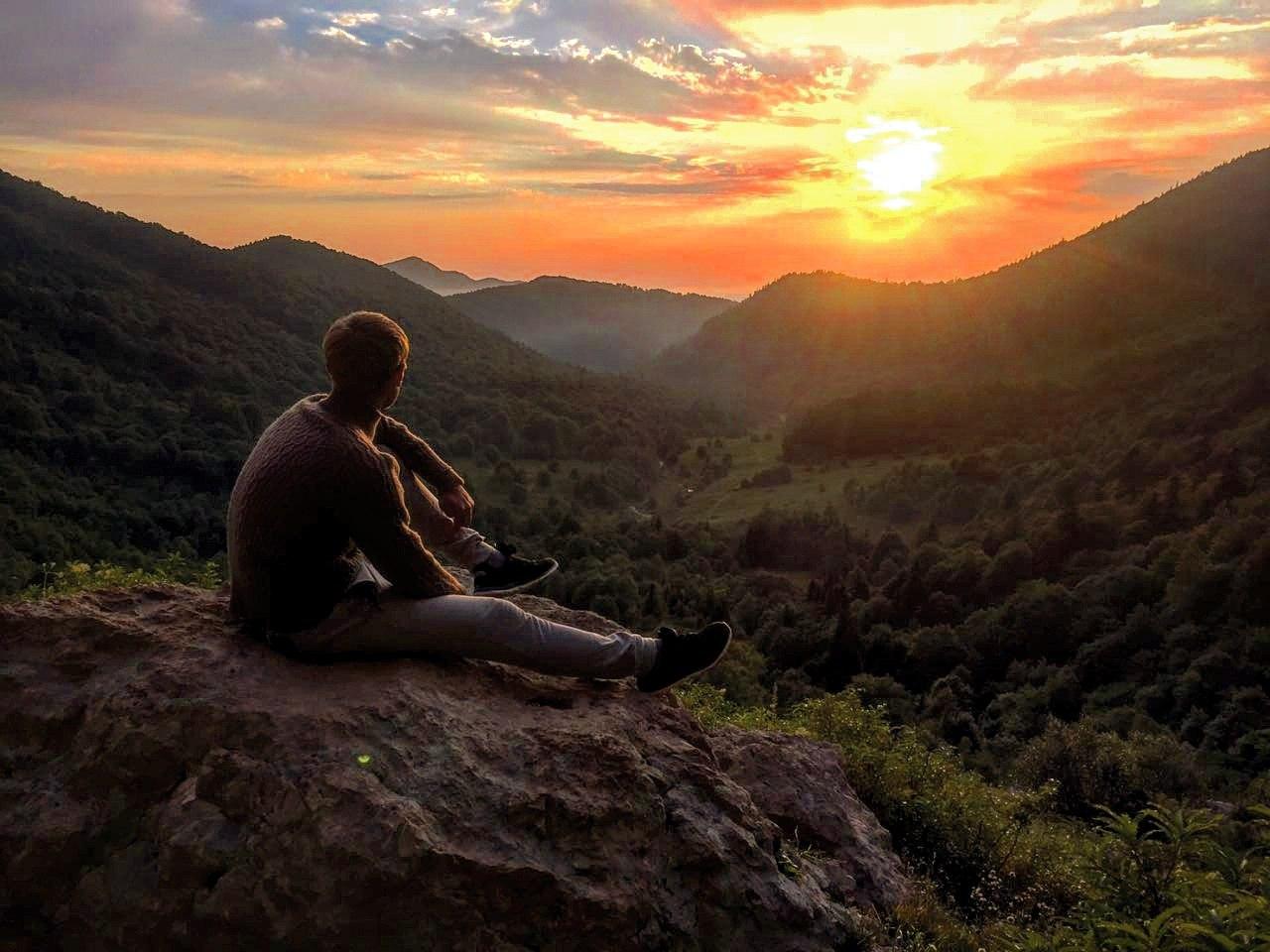 Молодой человек сидит на камне и фотографируется на фоне заката высоко в горах, природа красивая