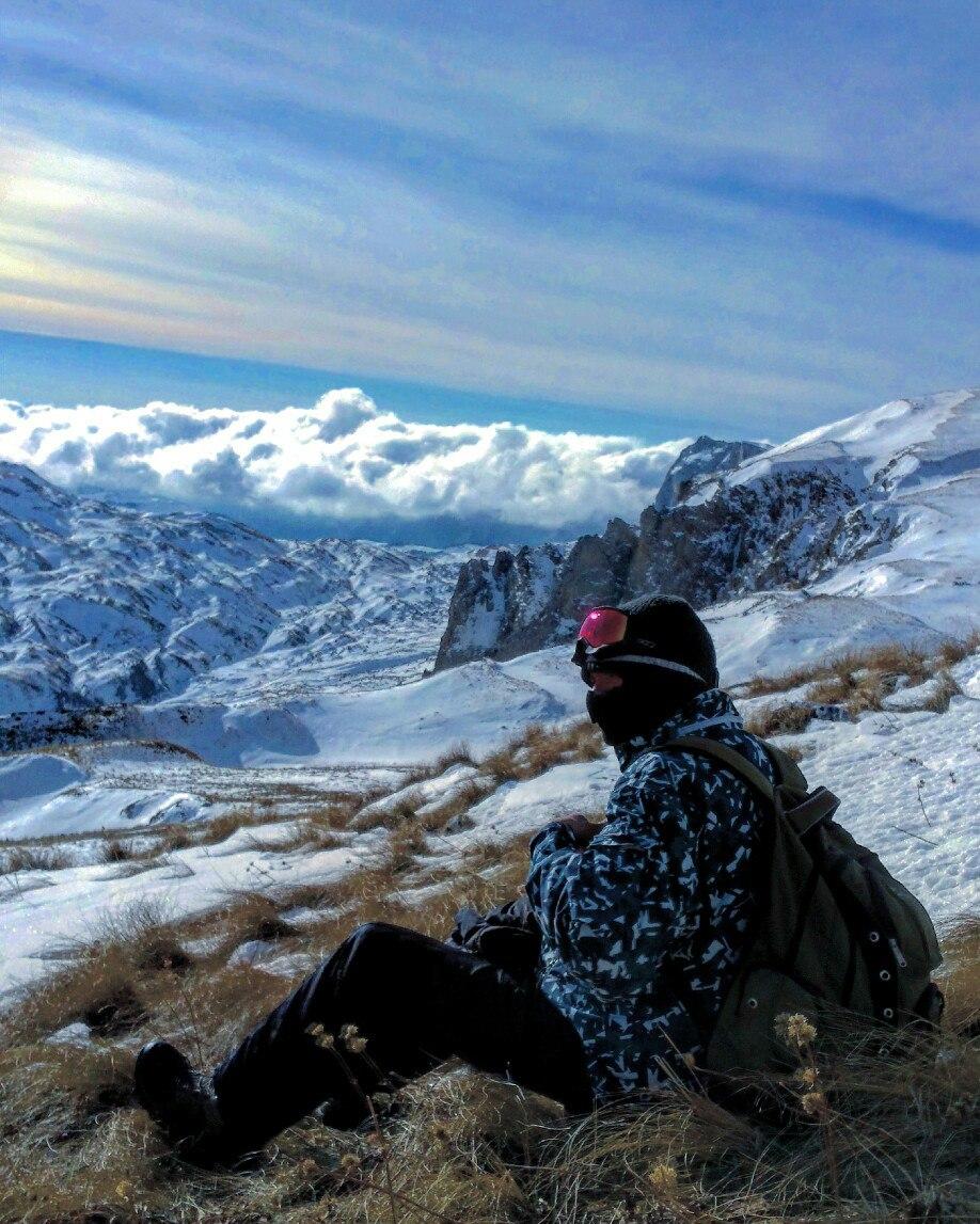 Молодой человек высоко в снежных горах сидит и отдыхает в маске и горнолыжных очках, на спине рюкзак